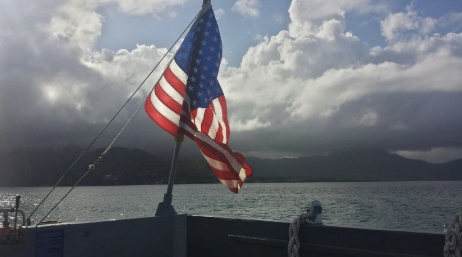 An American flag flies at the back of a boat, in water, overlooking hills in the distance on a cloudy day.