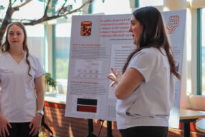 Two women are next to a presentation poster, actively discussing its content at a professional gathering.