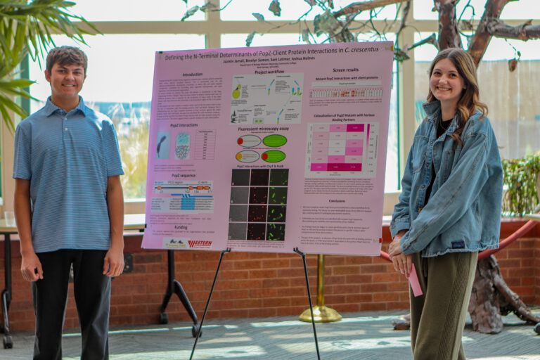 Two smiling students, one male and one female, stand beside a pink poster, showcasing their project