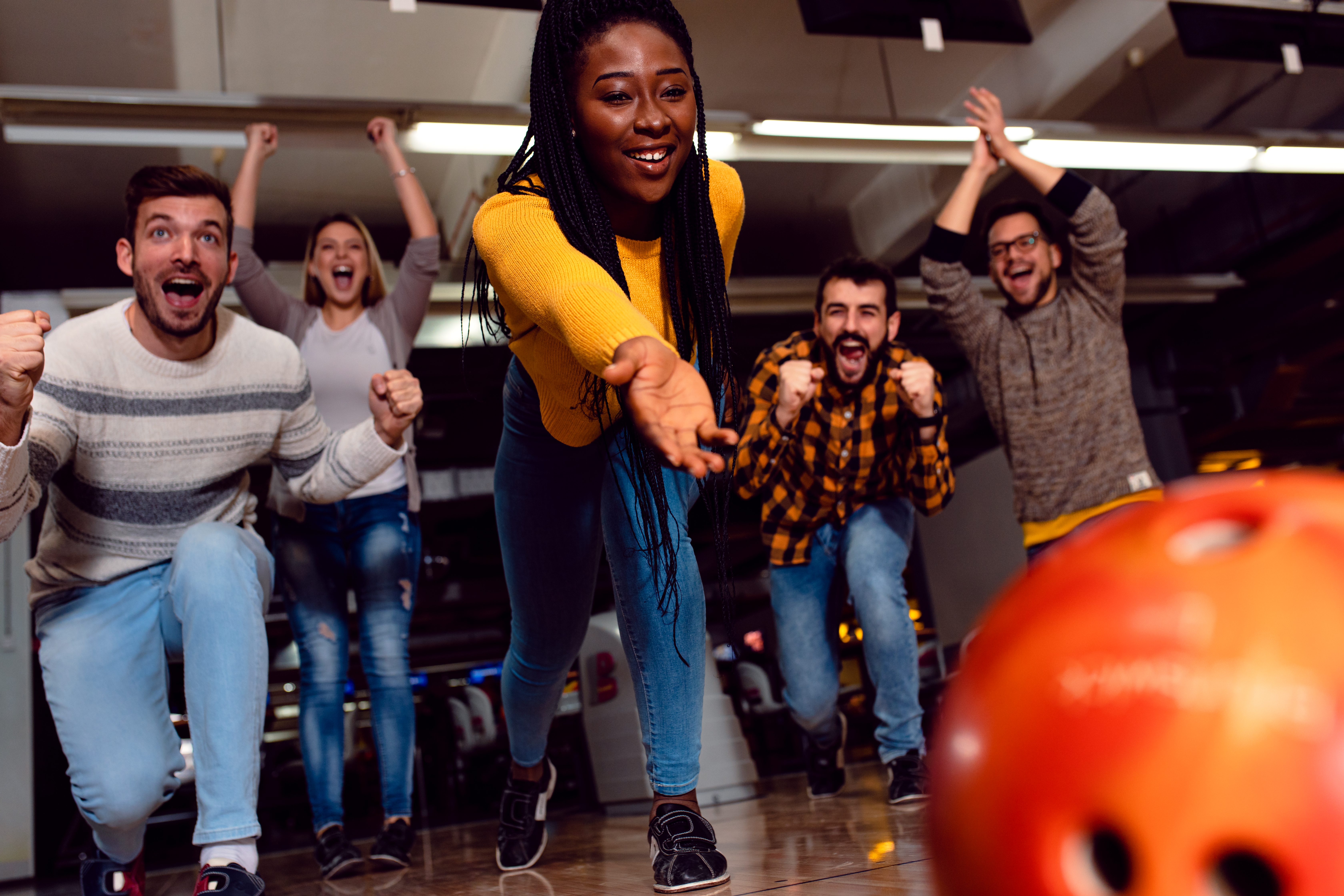 Diverse group of friends cheering one woman on as she releases an orange bowling ball