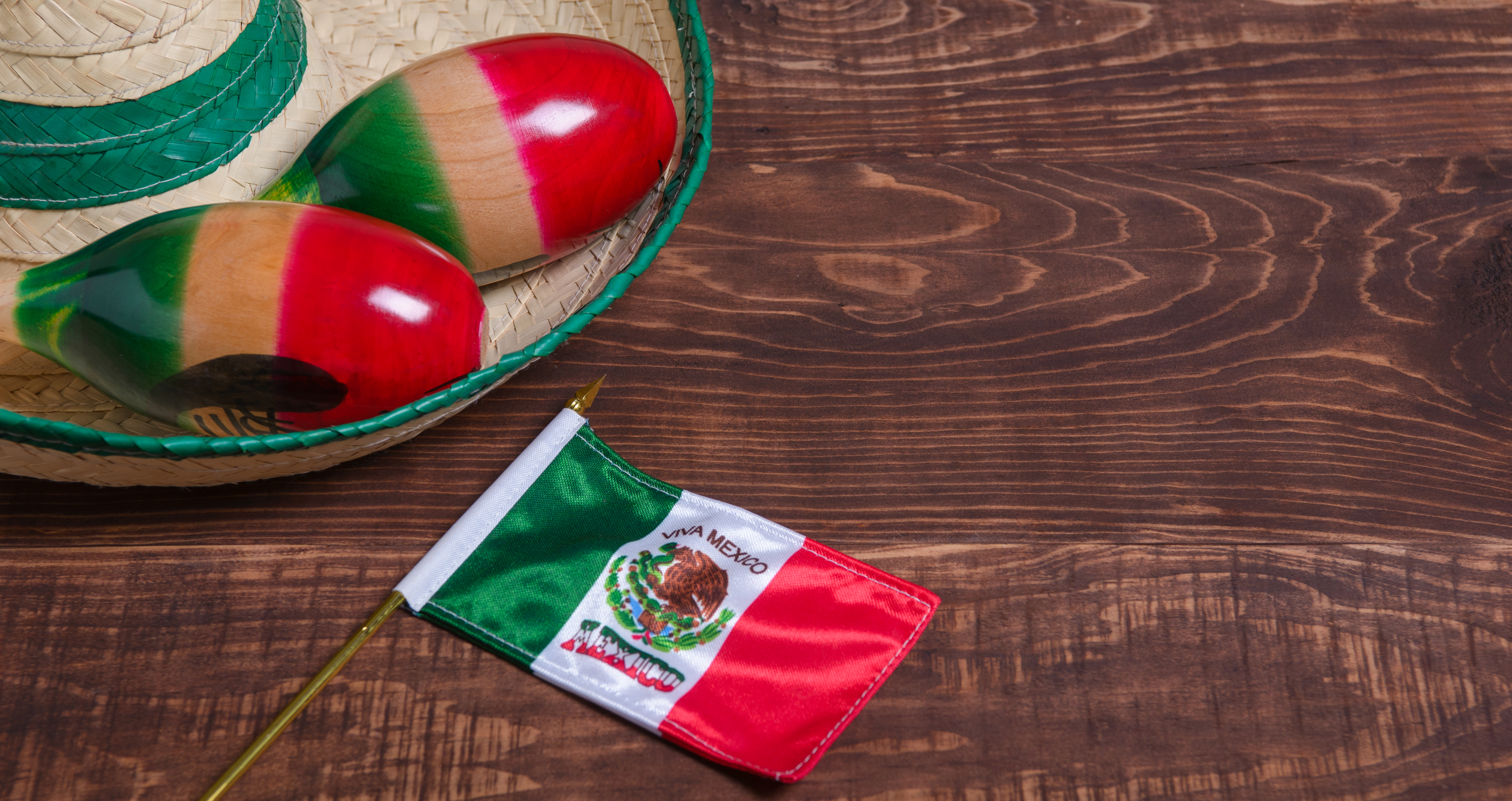 A small Mexican flag lies on a dark wooden surface next to a straw sombrero with green trim. Inside the hat sit a pair of maracas. The light is bright. The flag, hat, and maracas suggest a theme of celebration or national pride. 